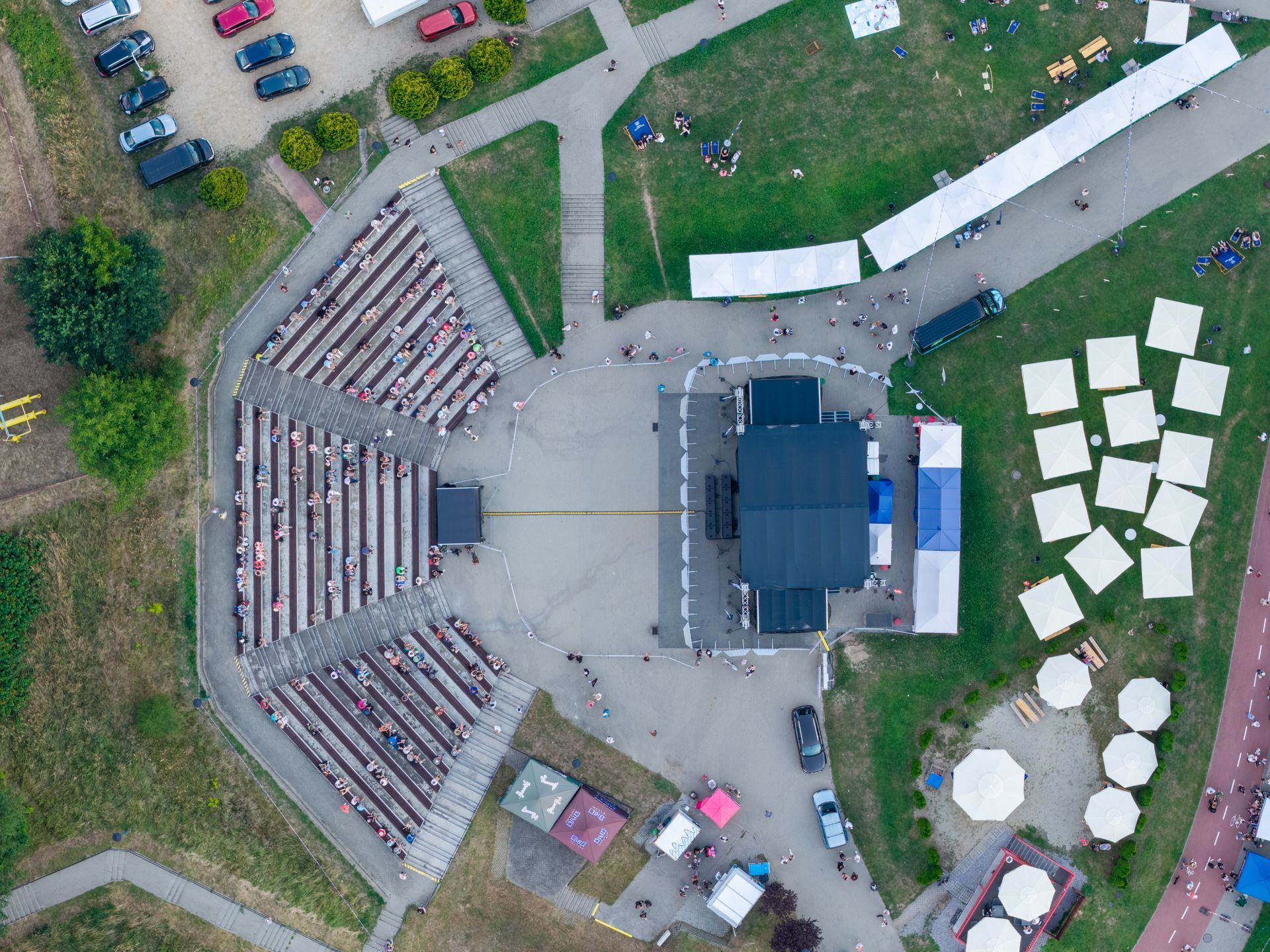 Vue aérienne d'un amphithéâtre en plein air avec des gradins incurvés, une scène centrale, et des tentes et voitures garées à proximité.