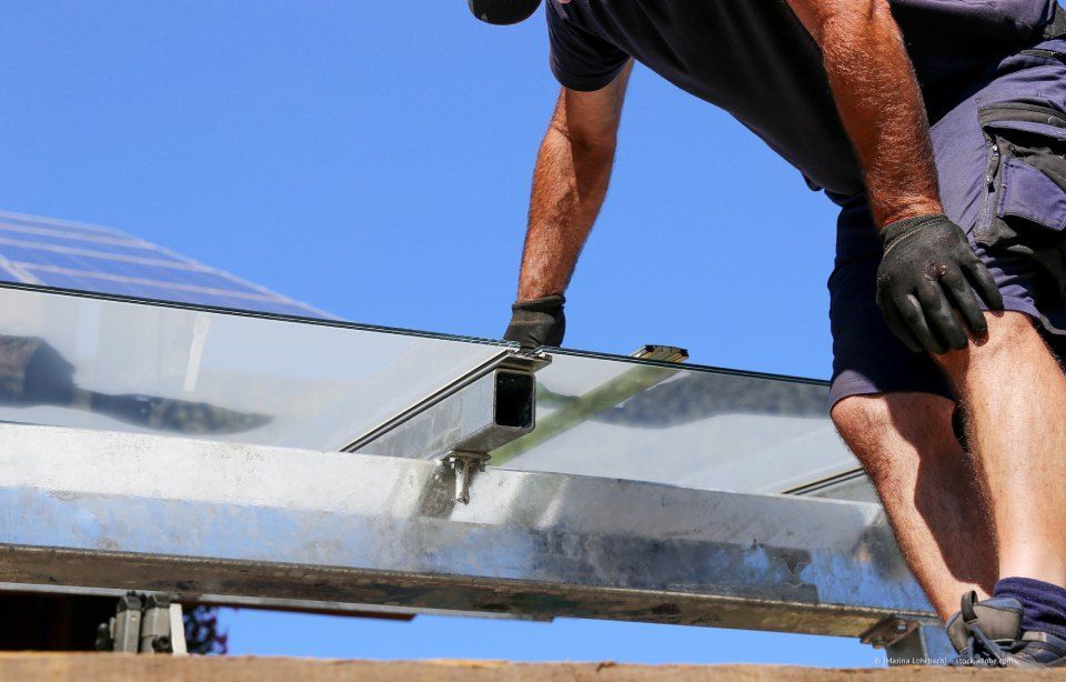 Arbeiter mit Handschuhen trägt unter blauem Himmel Dichtmittel auf einen Metallträger auf einem Dach auf