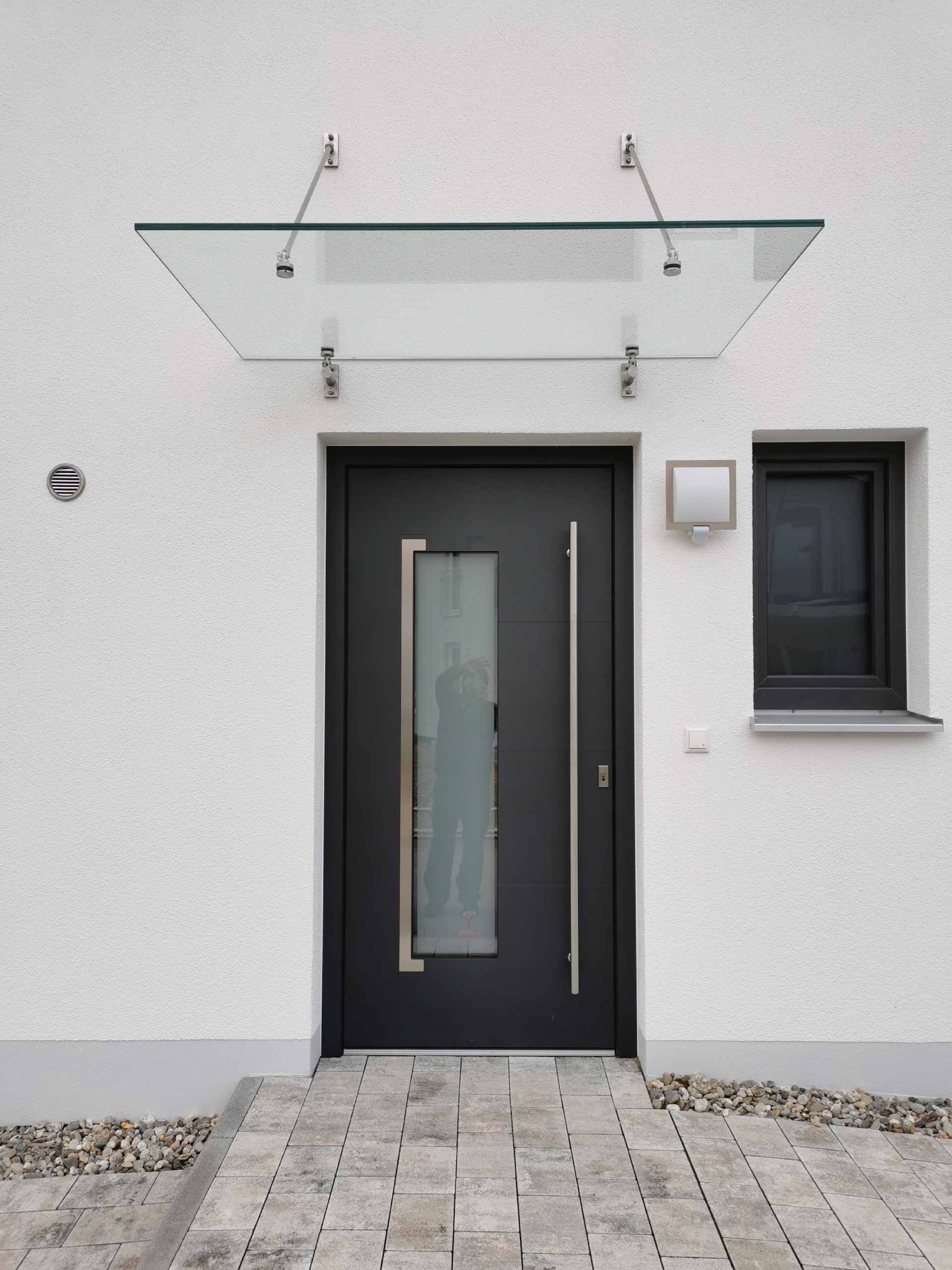 Moderner weißer Hauseingang mit dunkler Haustür, kleinem Fenster und Glasvordach über dem Eingang.