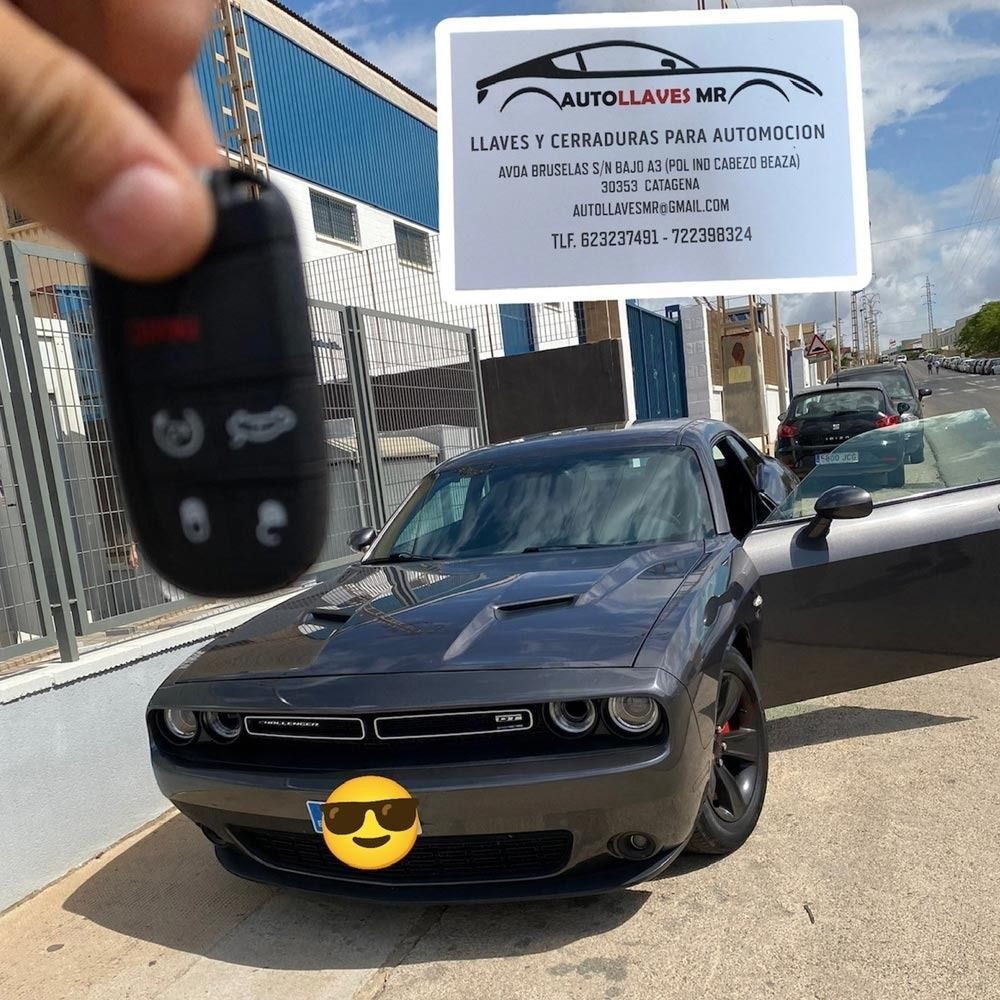 Persona con llave de coche junto a un Dodge Challenger gris. Tarjeta de visita en la esquina superior derecha.