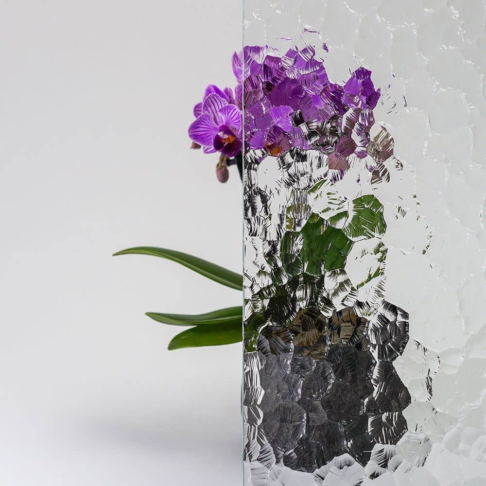 Lila Orchideen in einem dunklen Topf hinter strukturiertem Glas vor weißem Hintergrund.