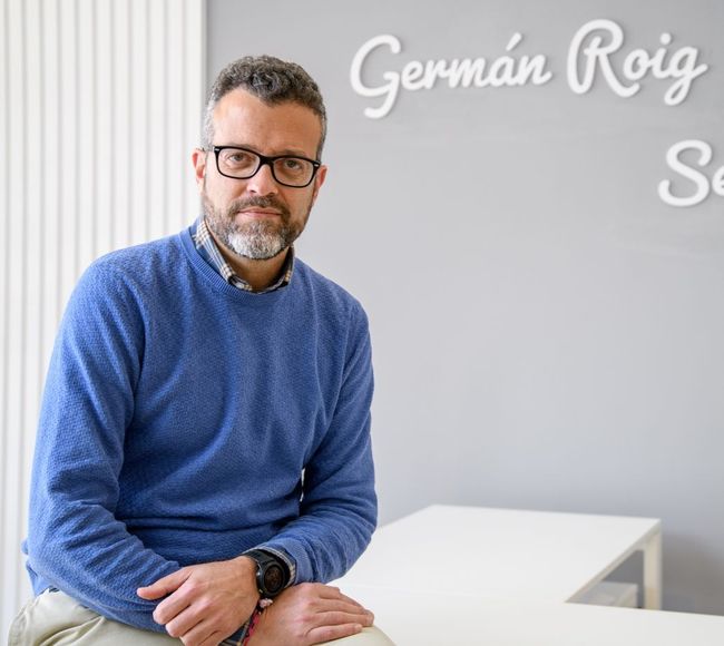 Un hombre con un suéter azul se sienta frente a una pared que dice Germán Roig.