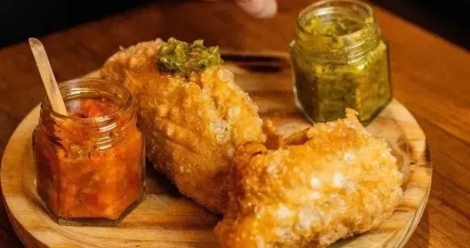 Dos empanadillas fritas crujientes sobre una tabla de madera con pequeños frascos de chutney rojo y verde.