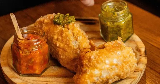 Dos empanadillas fritas crujientes sobre una tabla de madera con pequeños frascos de chutney rojo y verde.