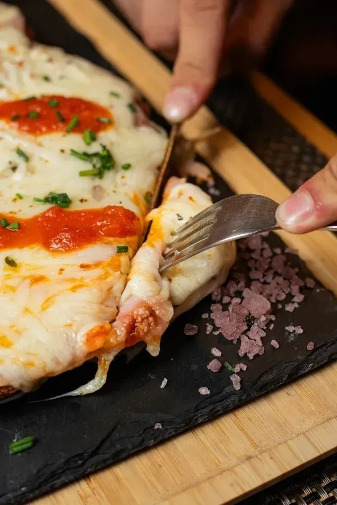 Manos cortando una porción de pizza de queso con tomate sobre una tabla de pizarra negra.