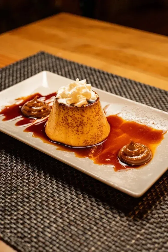 Postre de flan de crema pastelera con nata montada y salsa de caramelo en un plato blanco, sobre un camino de mesa.