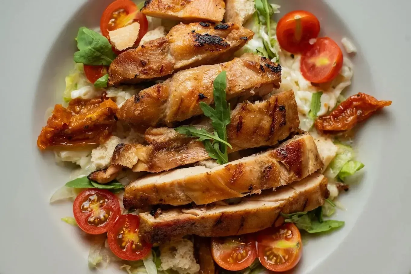 Lonchas de pollo a la parrilla en un plato blanco con tomates cherry, lechuga y hierbas aromáticas.
