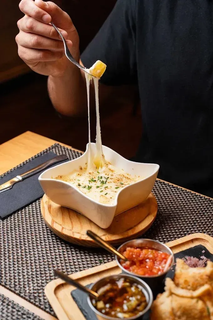 Una persona saca queso elástico de un tazón de salsa cremosa con salsas aparte sobre una mesa de madera.