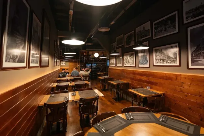 Comedor vacío de un restaurante con paredes de madera, fotos enmarcadas y mesas pequeñas bajo luces cálidas del techo.