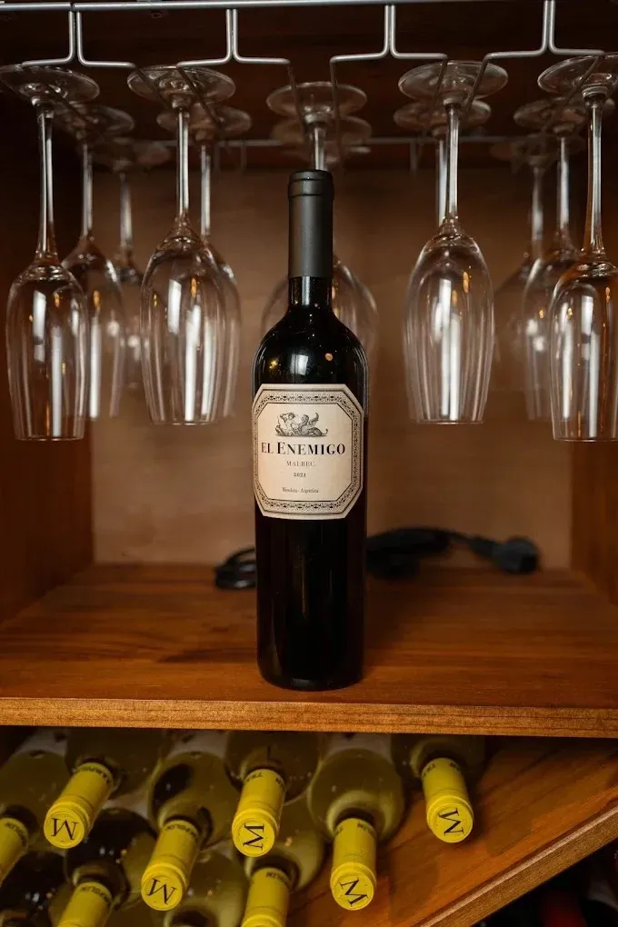 Botella de vino centrada bajo copas colgantes en un estante de madera con otras botellas debajo.