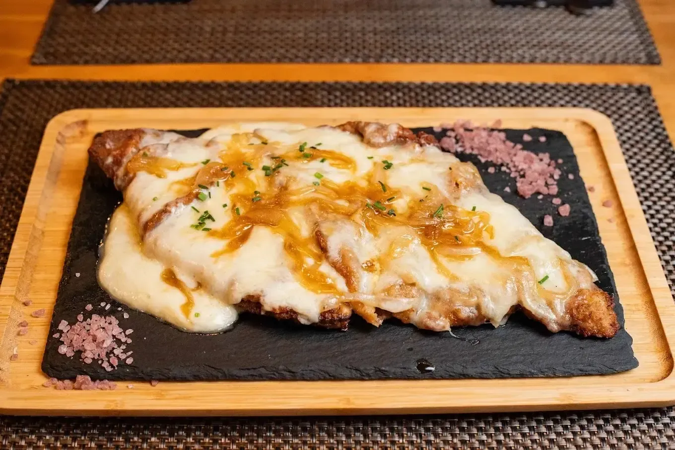Plato gratinado con queso sobre un plato de pizarra negra con hierbas, servido sobre un mantel individual tejido.