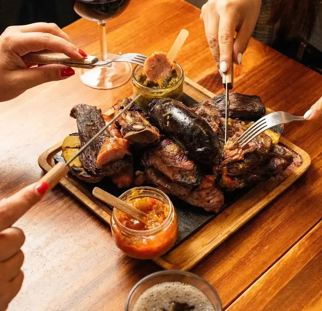 Manos que utilizan utensilios para compartir carne a la parrilla con salsas para mojar en una mesa de madera.