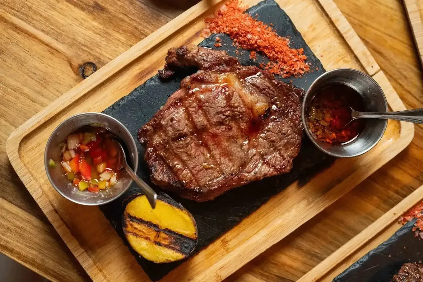 Filete a la parrilla en un plato negro con salsas y patatas asadas en una bandeja de madera.