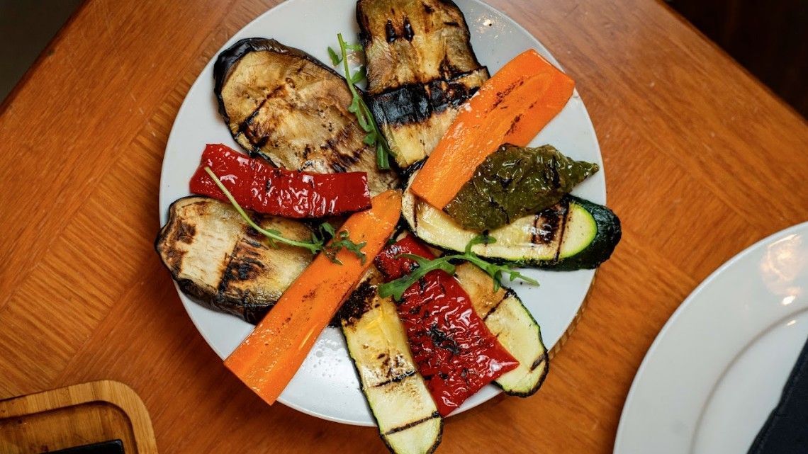 Mezcla de verduras a la parrilla en un plato blanco: calabacín, berenjena, pimiento rojo y zanahorias en una mesa de madera.
