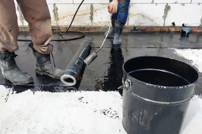 Trabajadores aplican membrana negra para techos con un soplete, usando un balde de asfalto líquido.