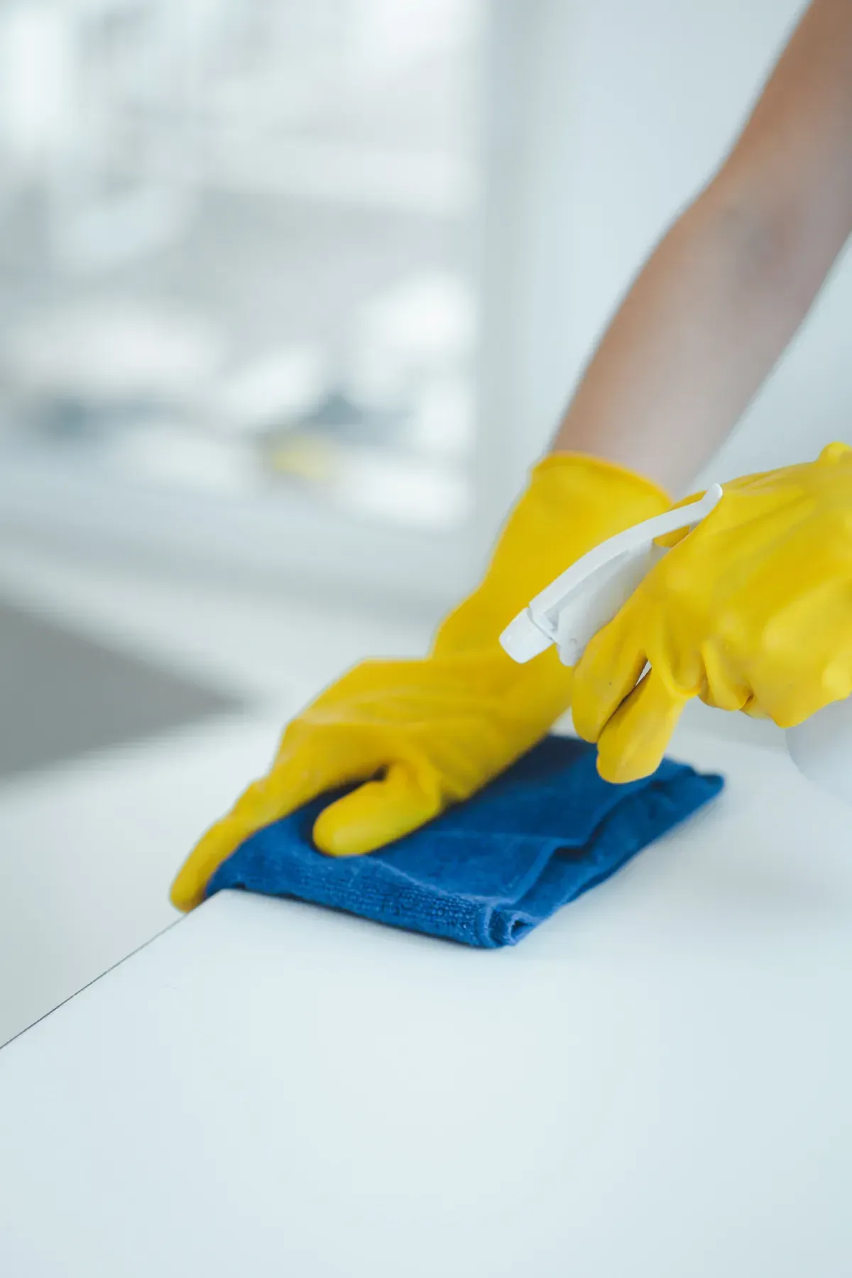 Persona que usa guantes amarillos limpiando una superficie blanca con un paño azul y una botella rociadora.