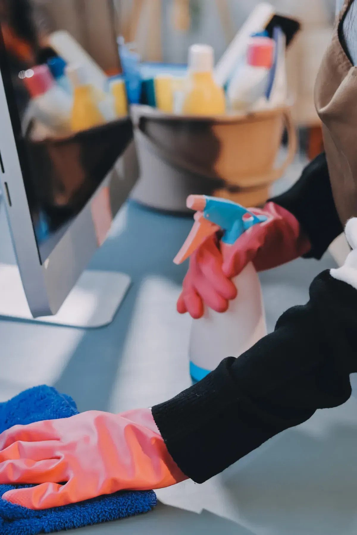 Una persona que usa guantes rosas rocía y limpia un escritorio con productos de limpieza y un paño azul.