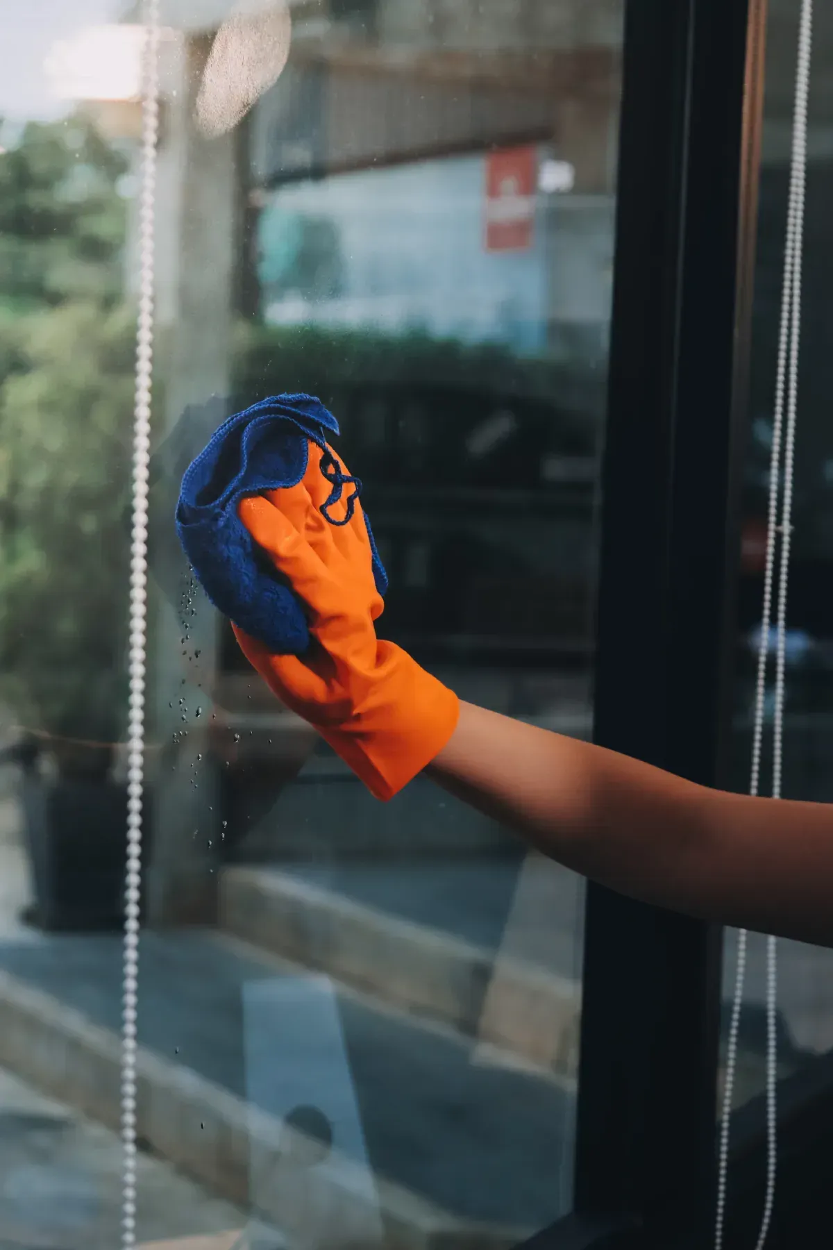 Mano con guante naranja limpiando una ventana con un paño azul.