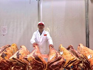 Un carnicero con bata y gorra blancas se encuentra frente a grandes cortes de carne colgados en una cámara refrigerada.