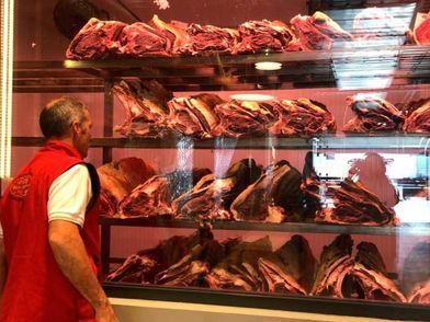 Carnicero de pie frente a una vitrina llena de diversos cortes de carne roja. Lleva un chaleco rojo.