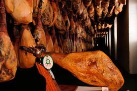 Piernas de jamón curado colgadas en una oscura cámara de almacenamiento; una en primer plano está lista para ser cortada.