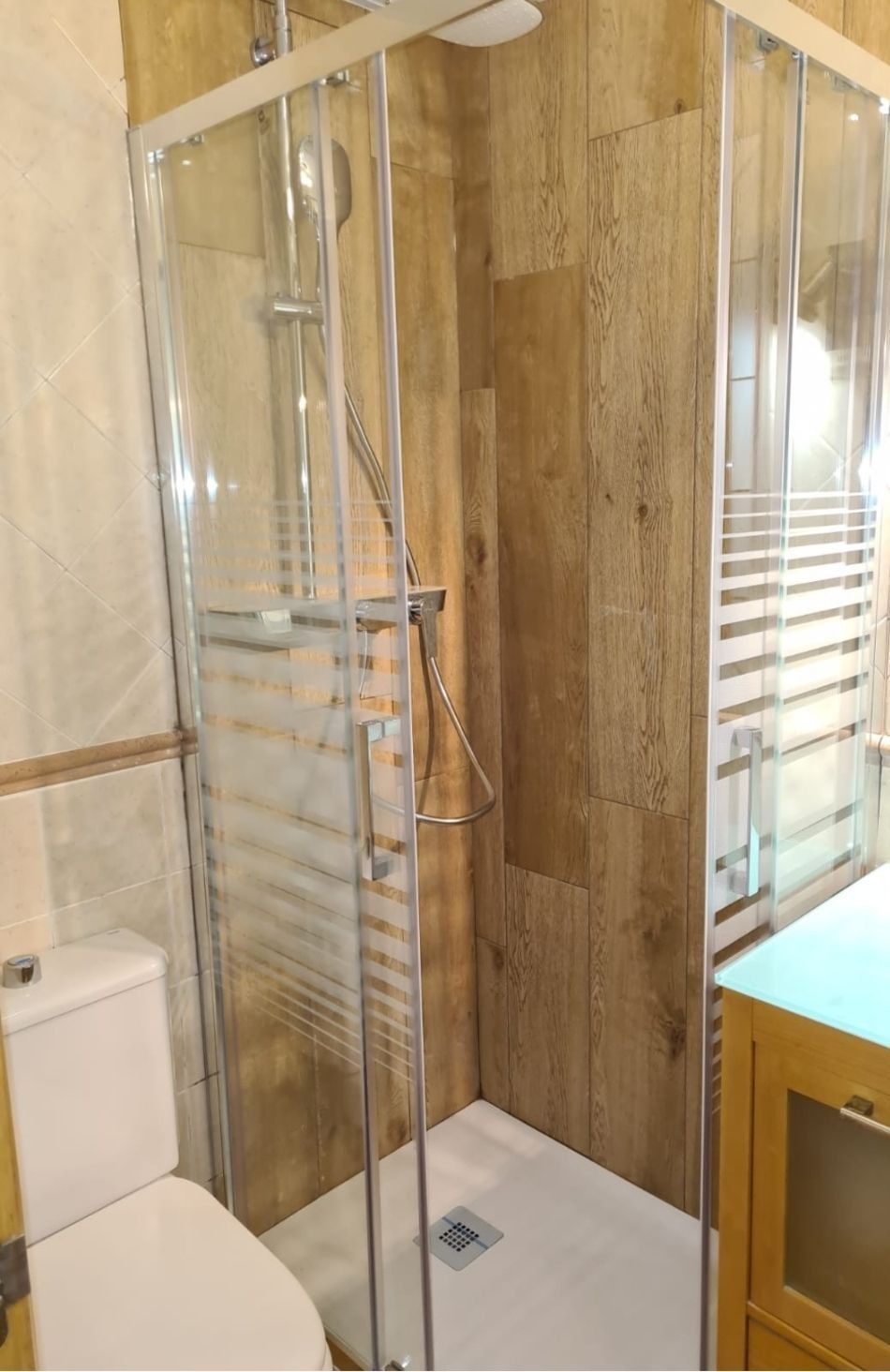 Baño pequeño con cabina de ducha de cristal, revestimiento de paredes con aspecto de madera y un inodoro blanco.
