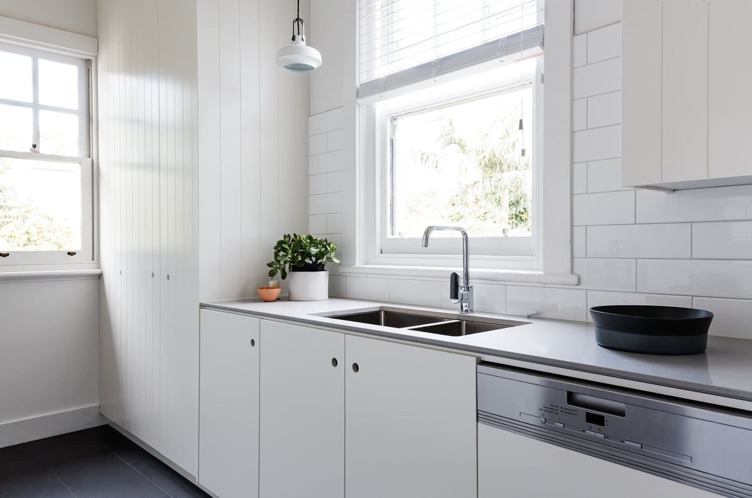 Cocina blanca luminosa con fregadero, ventana, armarios y una planta en maceta sobre la encimera.