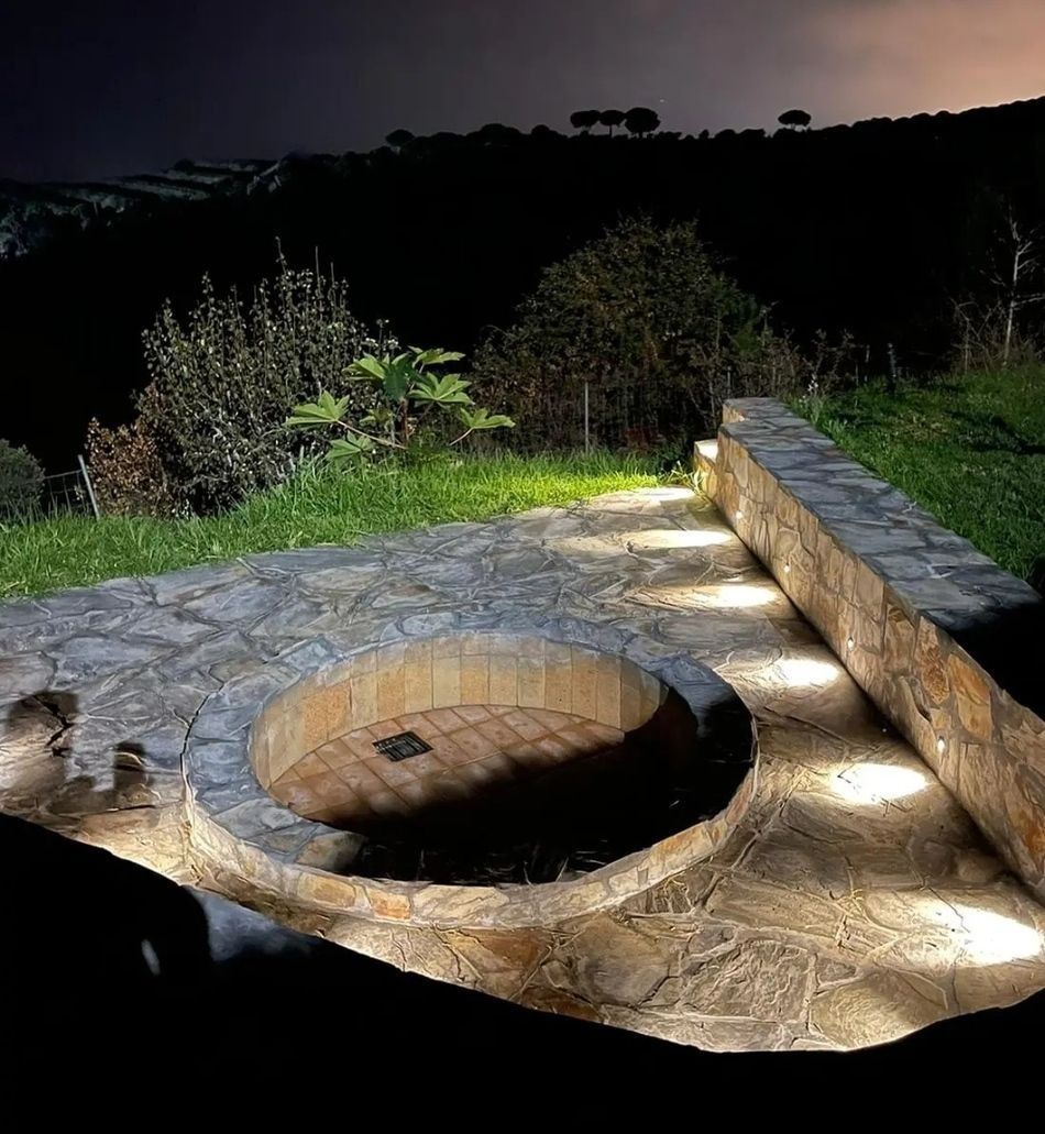 Hoguera de piedra con banco incorporado, iluminada por luces de suelo al anochecer en un jardín.