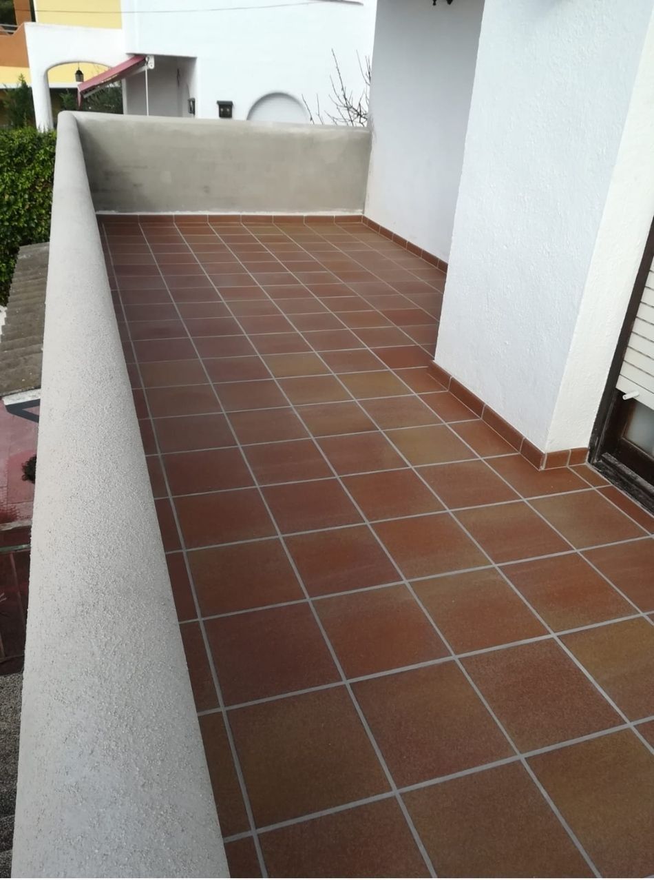 Estrecho pasillo de balcón con baldosas, paredes blancas y barandilla baja.