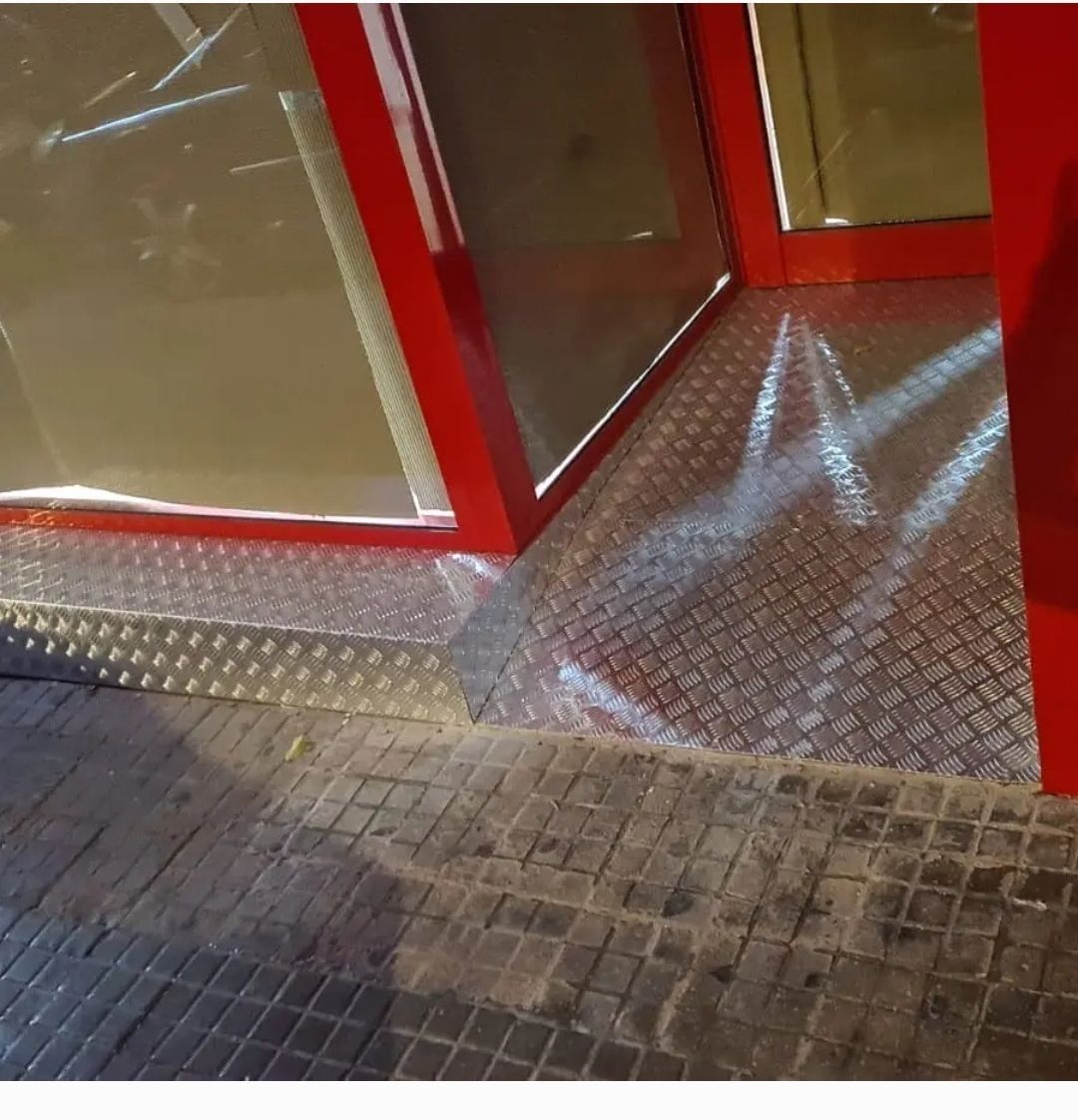 Entrada de cristal con marco rojo, rampa de chapa metálica antideslizante y pasarela de ladrillos en la parte delantera.