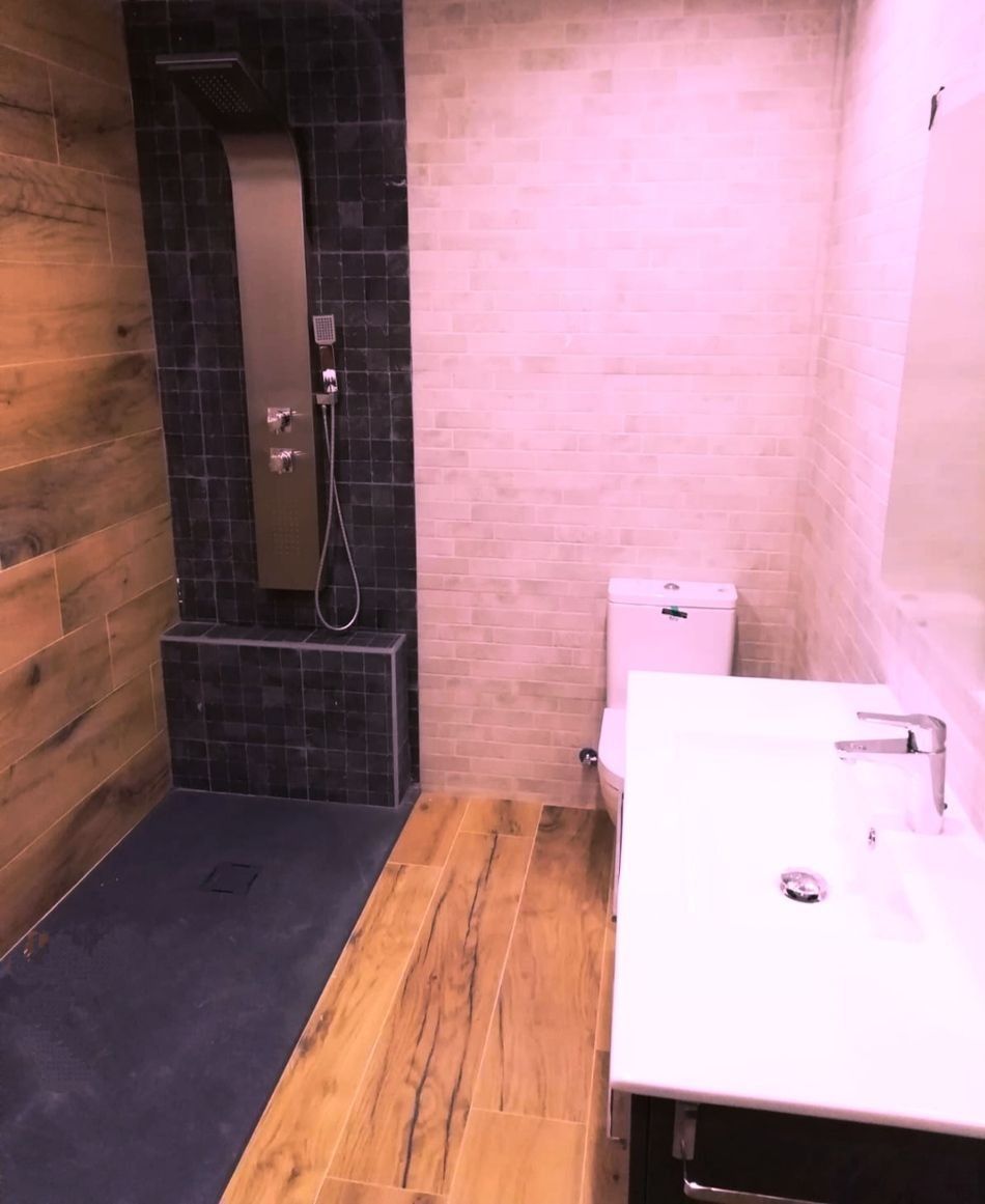 Baño moderno con ducha alicatada, suelo de imitación madera y lavabo e inodoro blancos.