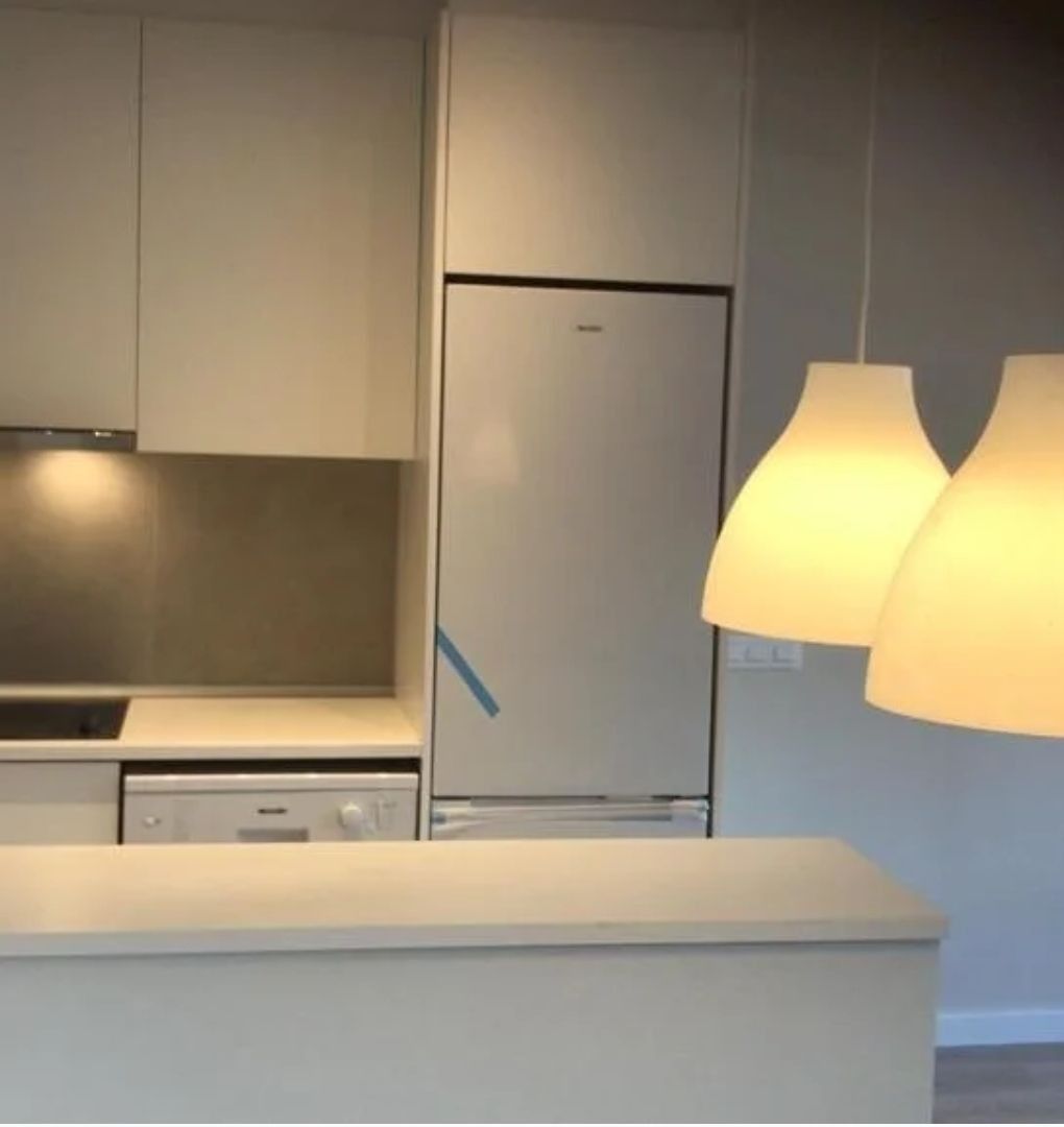 Cocina minimalista con armarios blancos, nevera de acero inoxidable y dos lámparas colgantes de luz cálida sobre la encimera.