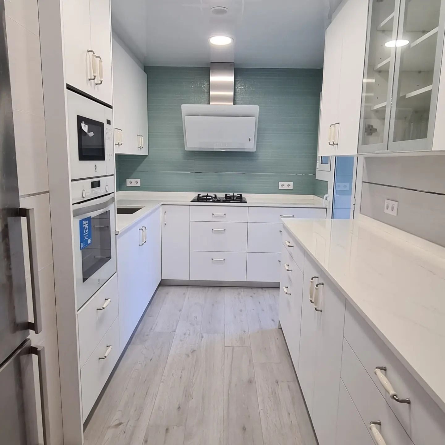 Cocina moderna blanca con salpicadero color turquesa, electrodomésticos de acero inoxidable y suelo de madera clara.