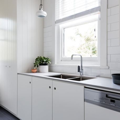 Cocina blanca luminosa con fregadero, ventana, armarios grises y pequeñas plantas en macetas.