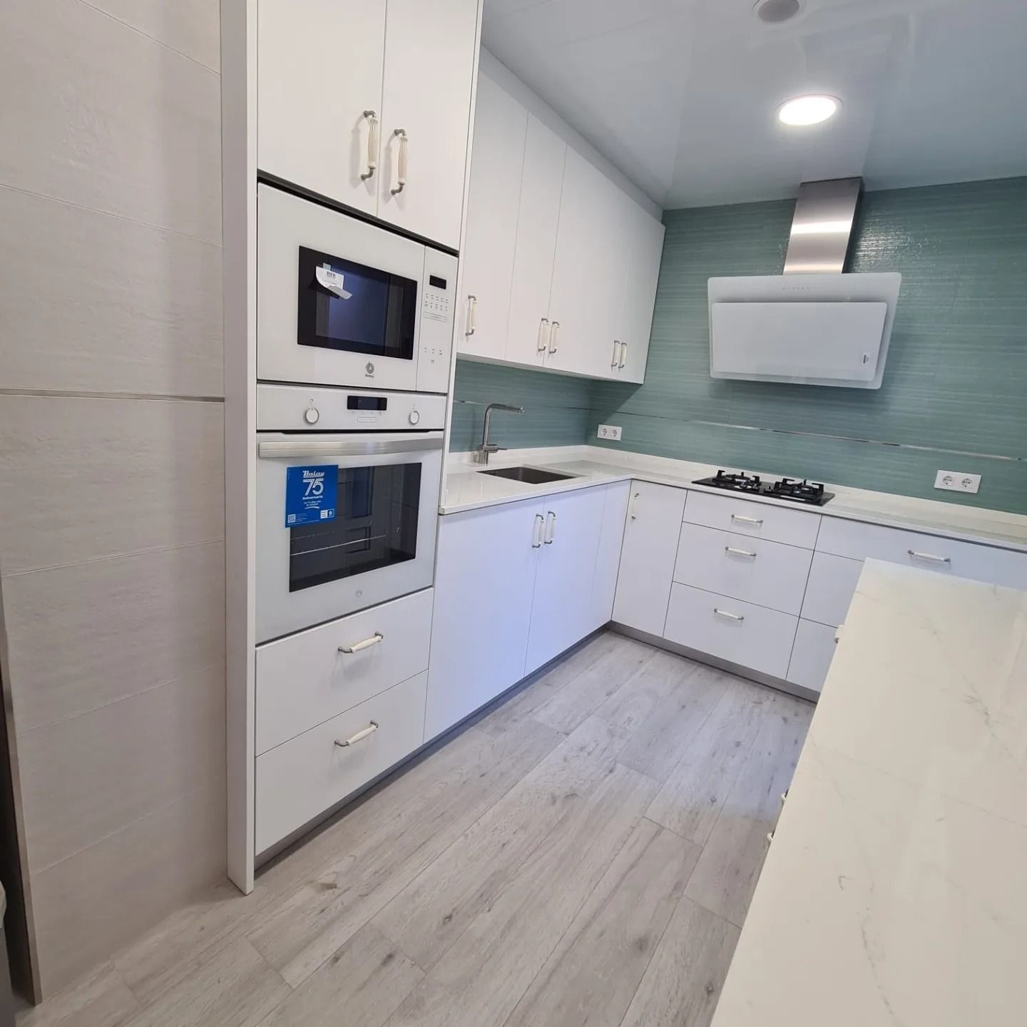 Cocina moderna blanca con hornos empotrados, salpicadero color turquesa y suelo de madera clara.