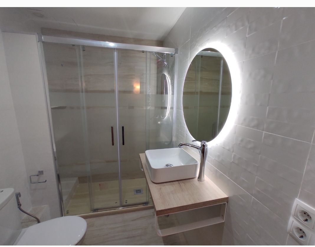 Baño moderno con ducha de cristal, espejo redondo retroiluminado y lavabo sobre encimera de madera.