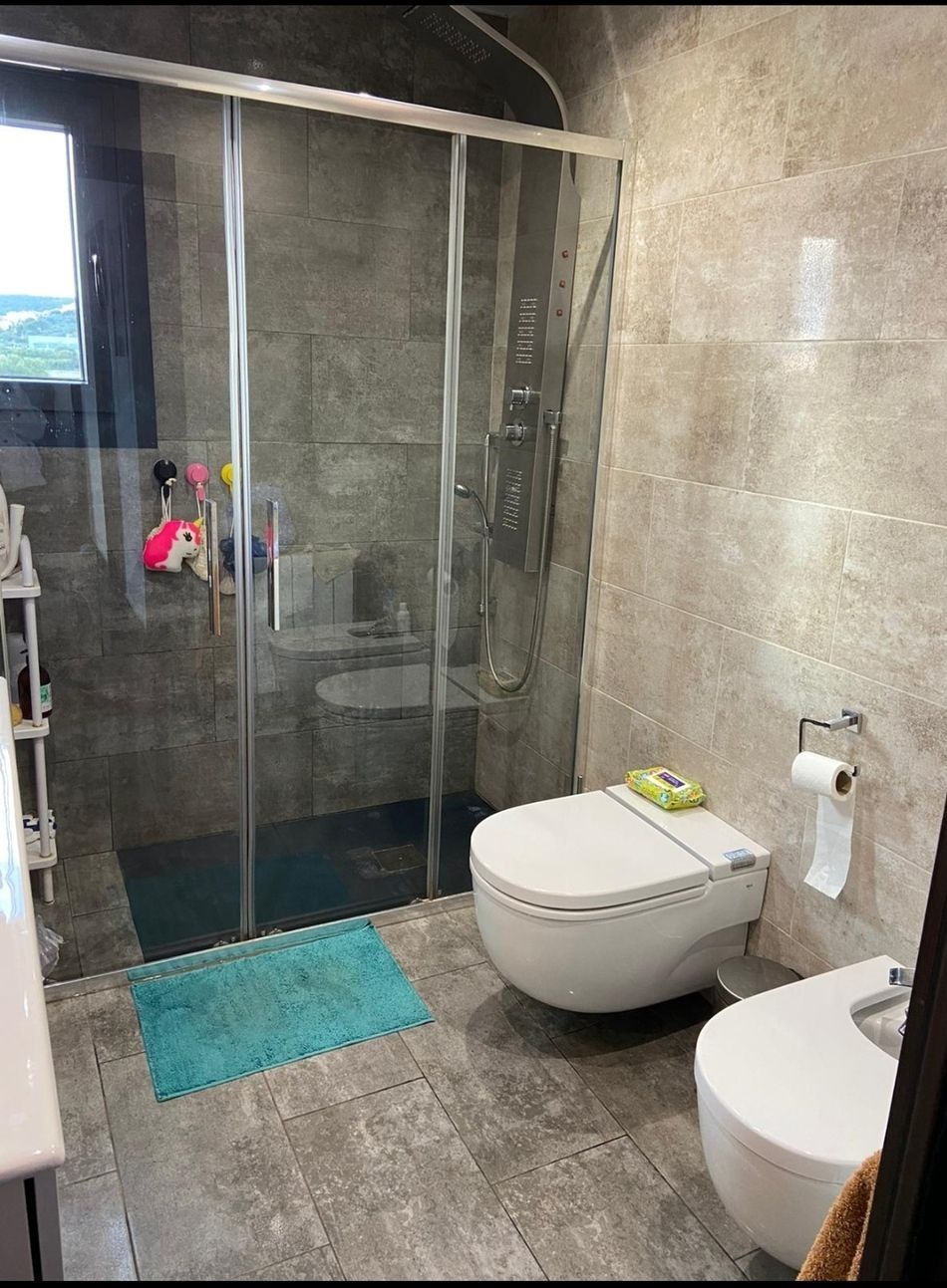 Baño moderno con ducha de cristal, inodoro, bidé y alfombrilla de baño azul sobre suelo de baldosas grises.
