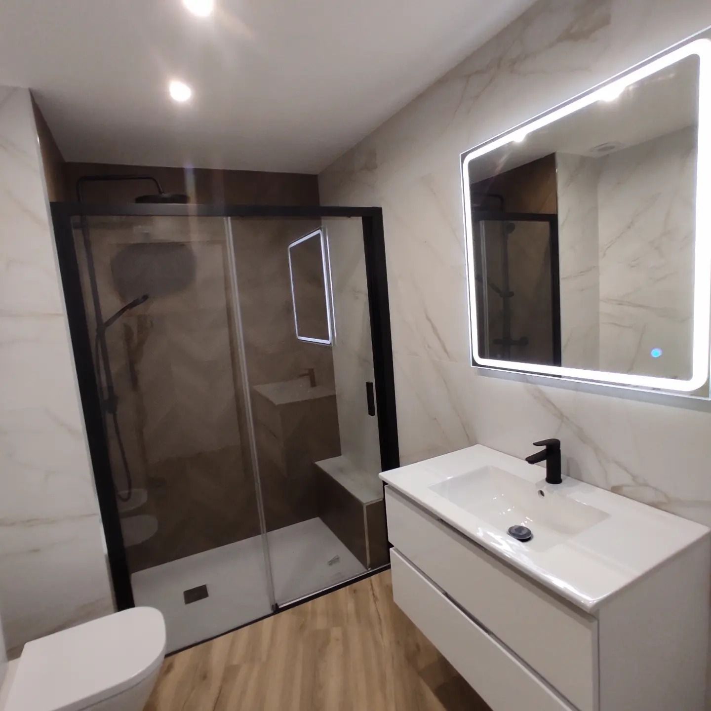 Baño moderno con ducha de cristal, lavabo suspendido, espejo iluminado y suelo con aspecto de madera.