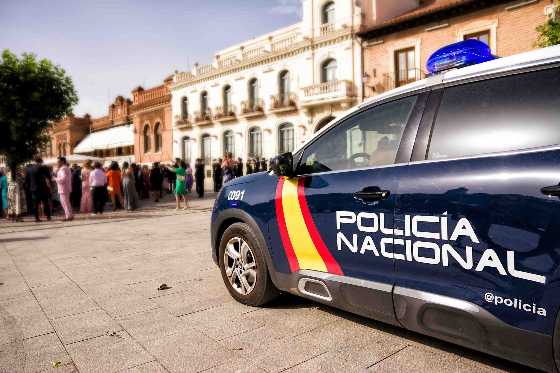 Coche de policía con los colores de la bandera española, aparcado sobre adoquines; grupo de personas al fondo.