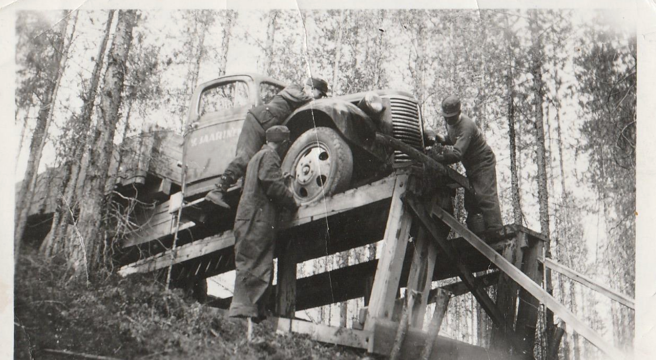 Mies seisoo puisella sillalla auton vieressä metsäalueella alhaalta päin katsottuna