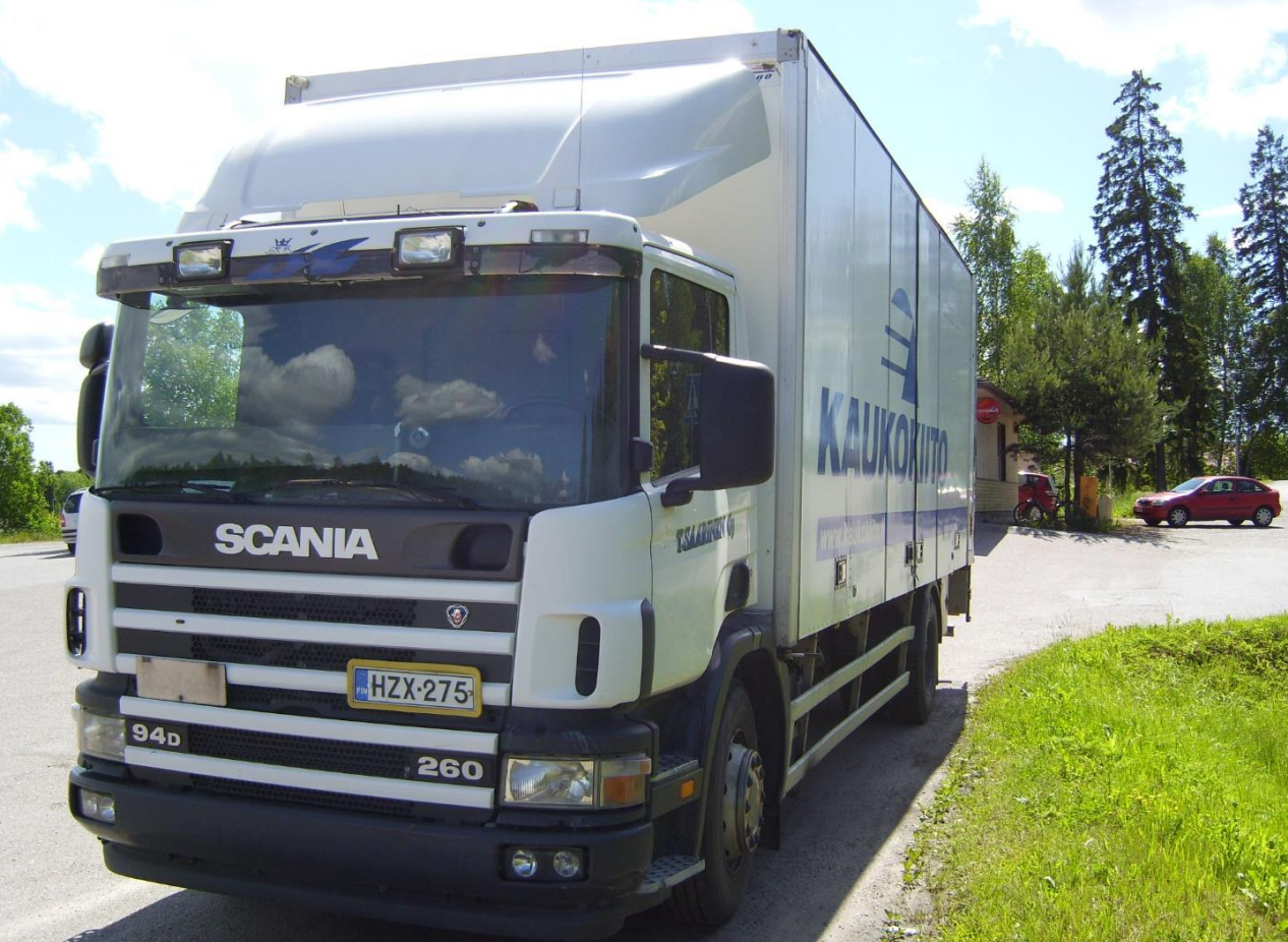 Valkoinen Scania-umpikorikuorma-auto pysäköitynä tien varteen puiden viereen, kirkas taivas ja punainen auto taustalla