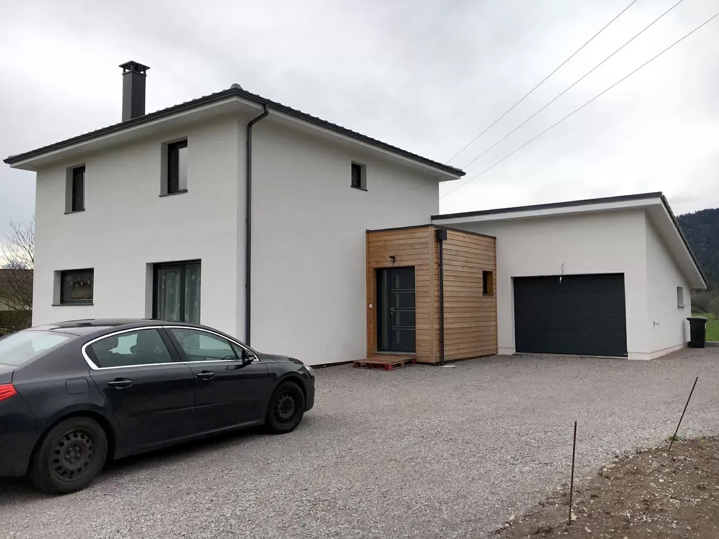 Maison blanche à deux étages avec une voiture noire garée sur une allée de gravier et un garage sur la droite.