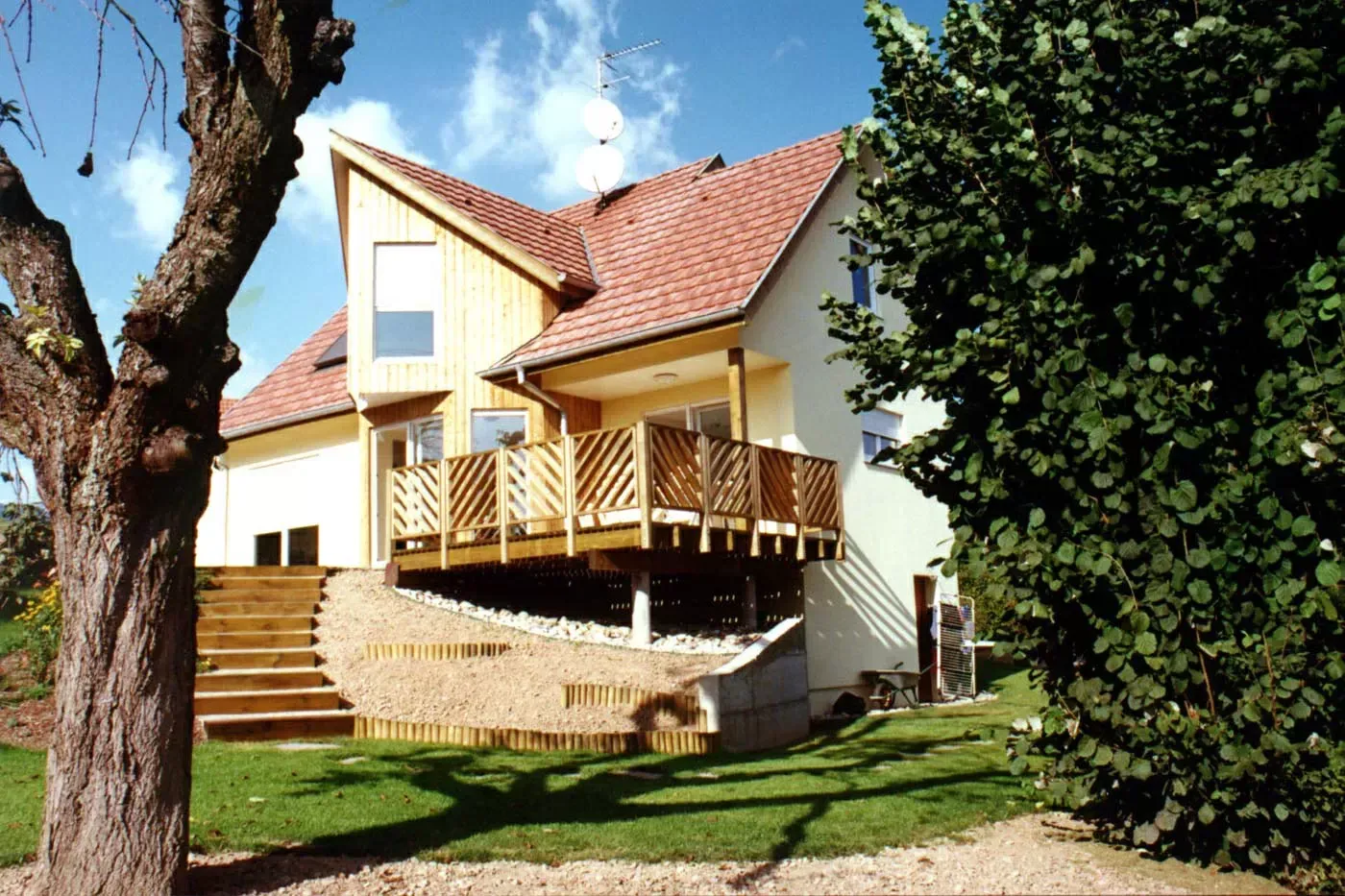 Une maison couleur crème avec un toit rouge, une terrasse en bois et de grands arbres dans un jardin ensoleillé