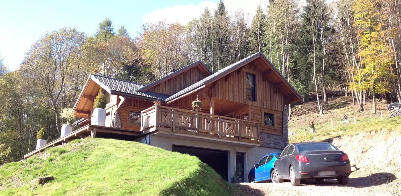 Chalet en bois sur une colline herbeuse, avec des voitures garées et des arbres d'automne en arrière-plan.