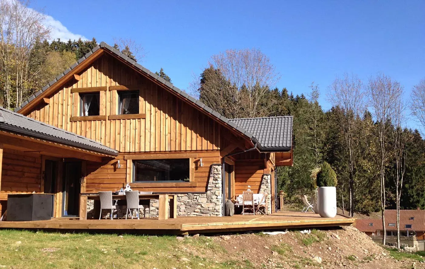 Cabane en bois avec soubassement en pierre et terrasse dans une clairière ensoleillée sous un ciel bleu