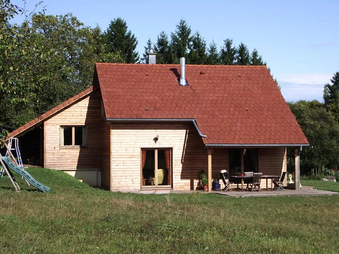 Petite maison beige au toit rouge, avec patio et jardin gazonné sous un ciel dégagé.