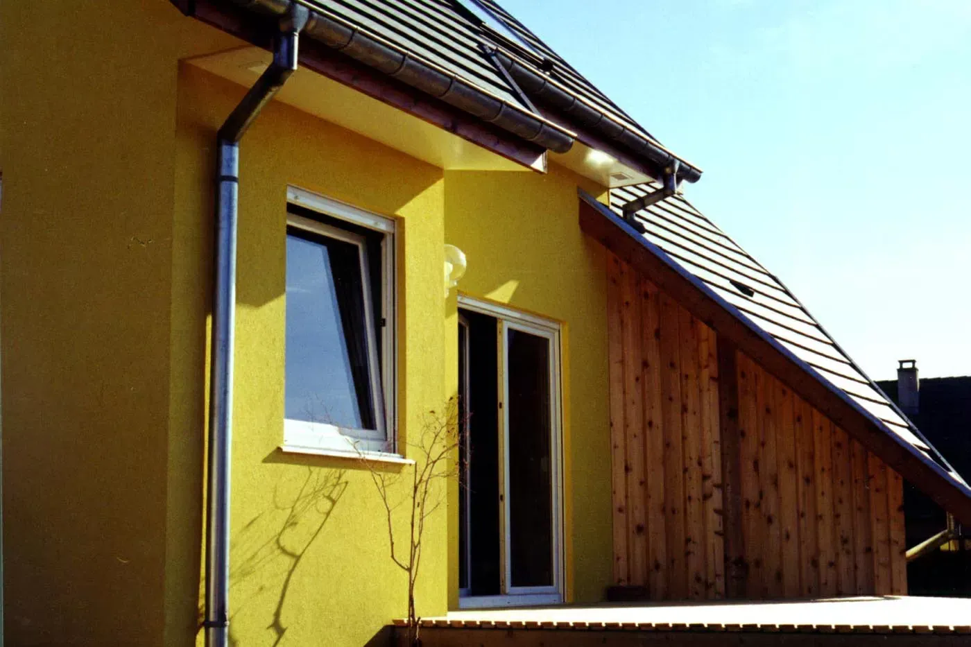 Maison jaune avec toit en pente et fenêtre ouverte à côté d'une extension revêtue de bois