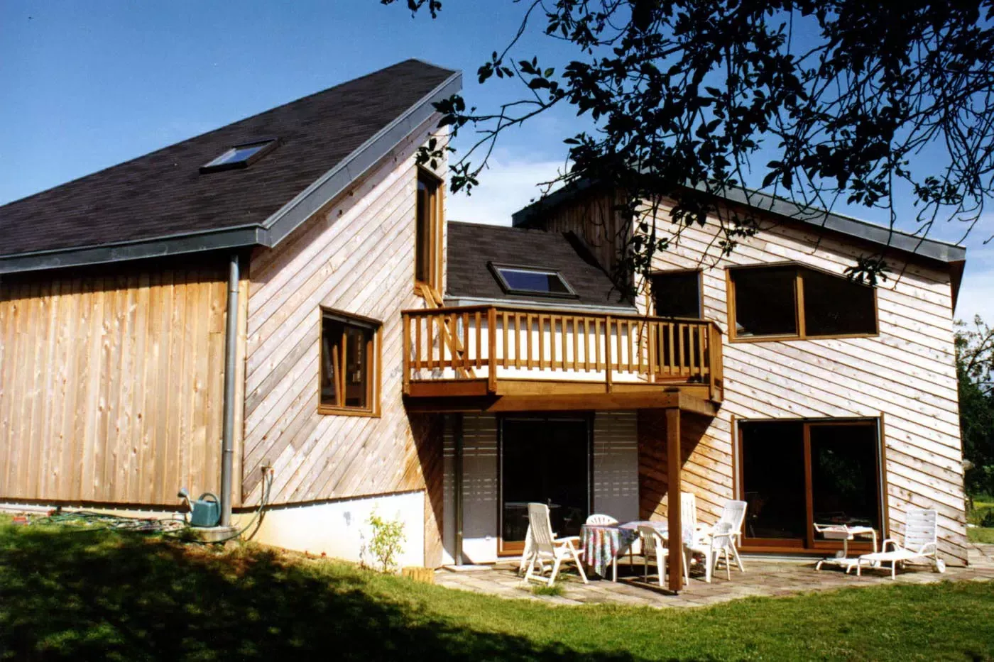 Maison en bois à deux étages avec balcon et chaises de patio dans une cour ensoleillée