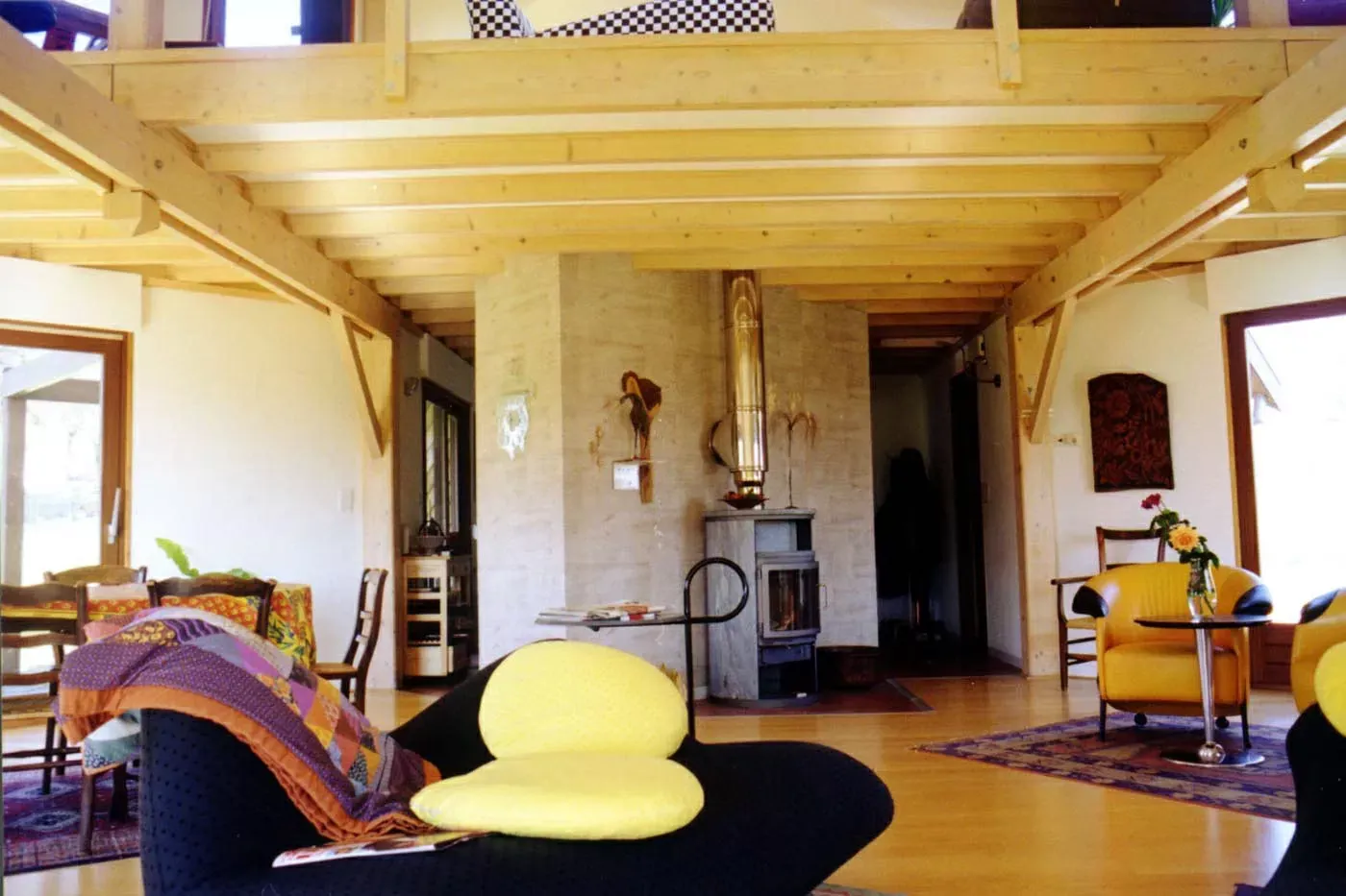 Salon chaleureux avec poutres apparentes, cheminée, chaises colorées et pouf jaune au premier plan.