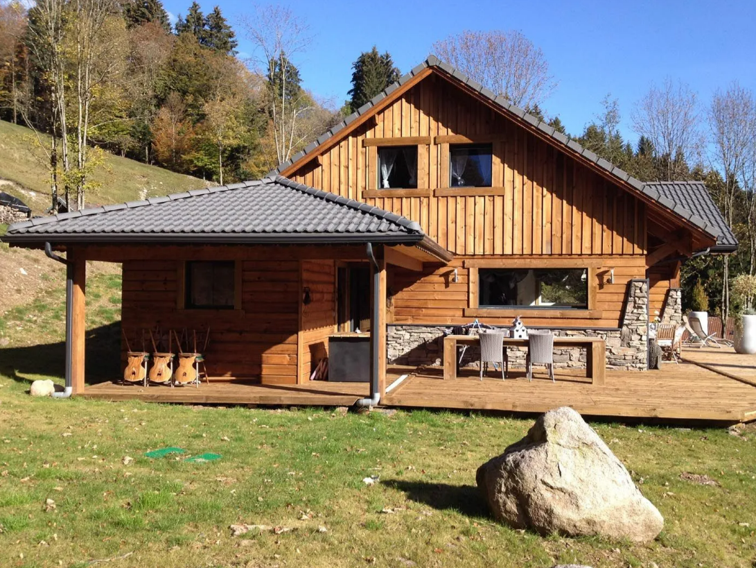 Cabane en bois avec toit gris et terrasse, située dans un cadre champêtre herbeux, avec un gros rocher devant.